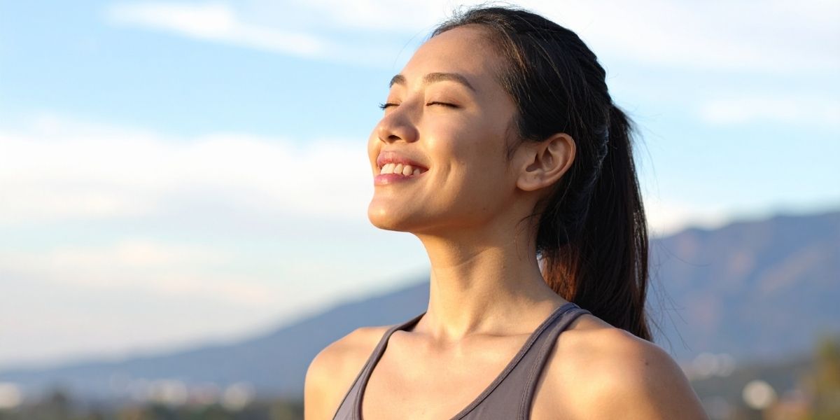 女性が目をつむり微笑んで立っている。表情はすがすがしく美しい。背景には遠くに山並みが広がっている。