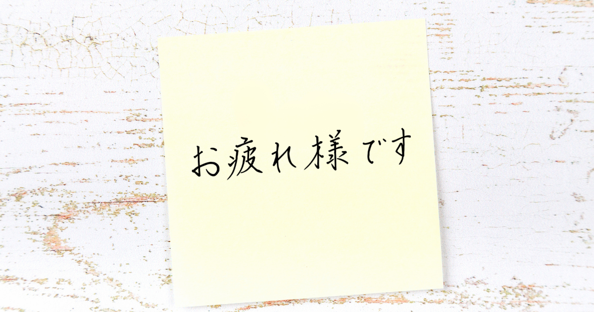 黄色い付箋に『お疲れさまです』とボールペンで手書きされた文字。白い木目の背景。